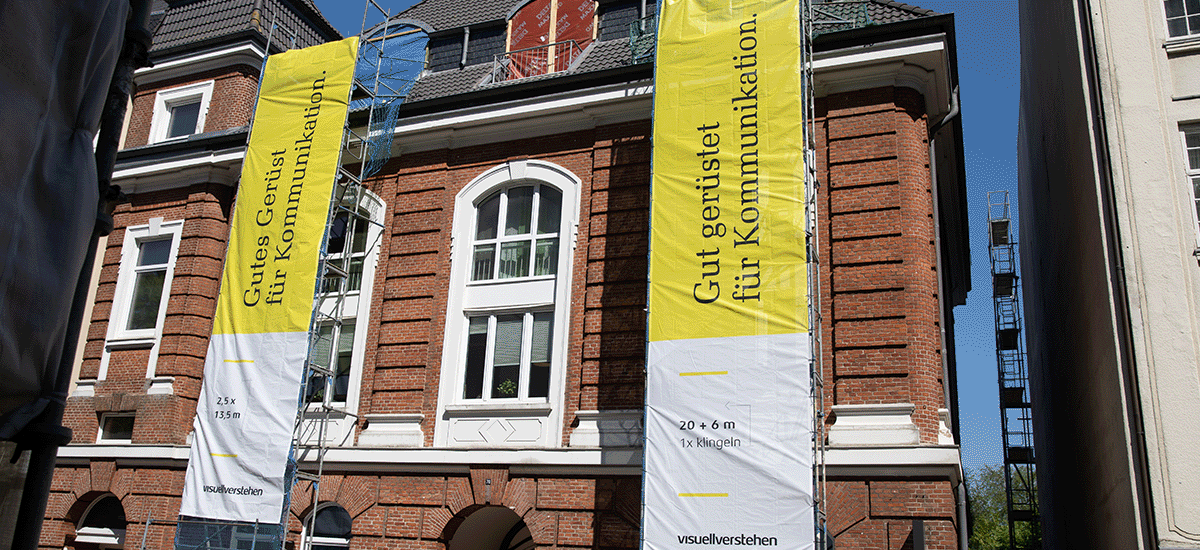 An einem Backsteingebäude hängt ein weiß-gelbes Bauzaunbanner mit schwarzer Schrift von visuellverstehen. Hinter dem Banner schaut ein junger Mann mit gelben Bauhelm hervor. Er präsentiert das Banner