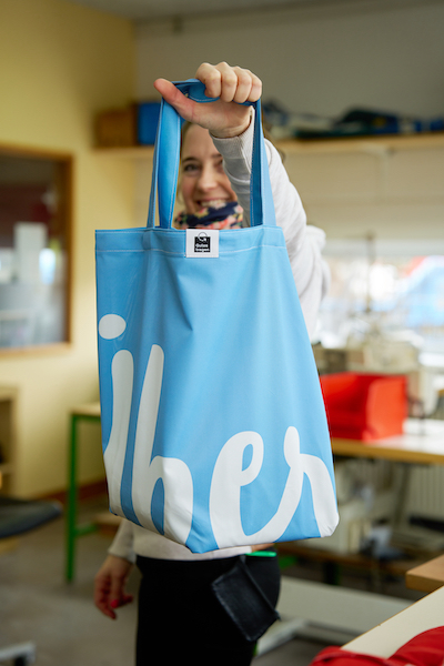 Ein Frau hält lächelnd eine blaue Tasche von Gutes Tragen hoch. Im Hintegrund sieht man die Werkstadt in der die Taschen hergestellt werden.