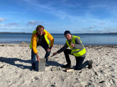 Zwei Mitglieder des Lions Club Flensburg knieen an einem Strand und öffnen eine Box aus Metall. Beide schauen in die Kamera, der eine Mann zeigt einen Daumen.