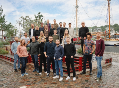 Ein Gruppenfoto von den Mitarbeiter:innen von visuellverstehen. Die 24 Männer und Frauen stehen vor und auf einer Bankkonstruktion am flensburger Hafen.